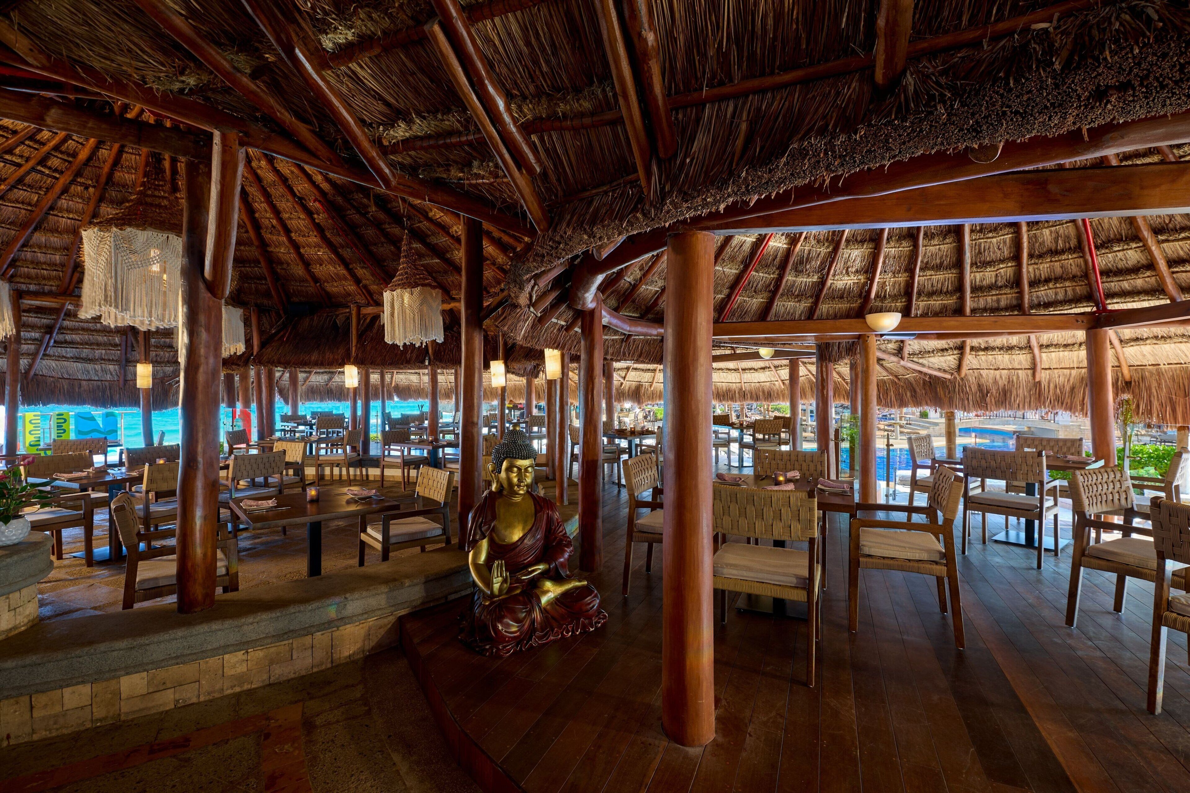 A spacious outdoor seating area with wooden tables and chairs, a Buddha statue, and a thatched roof structure.