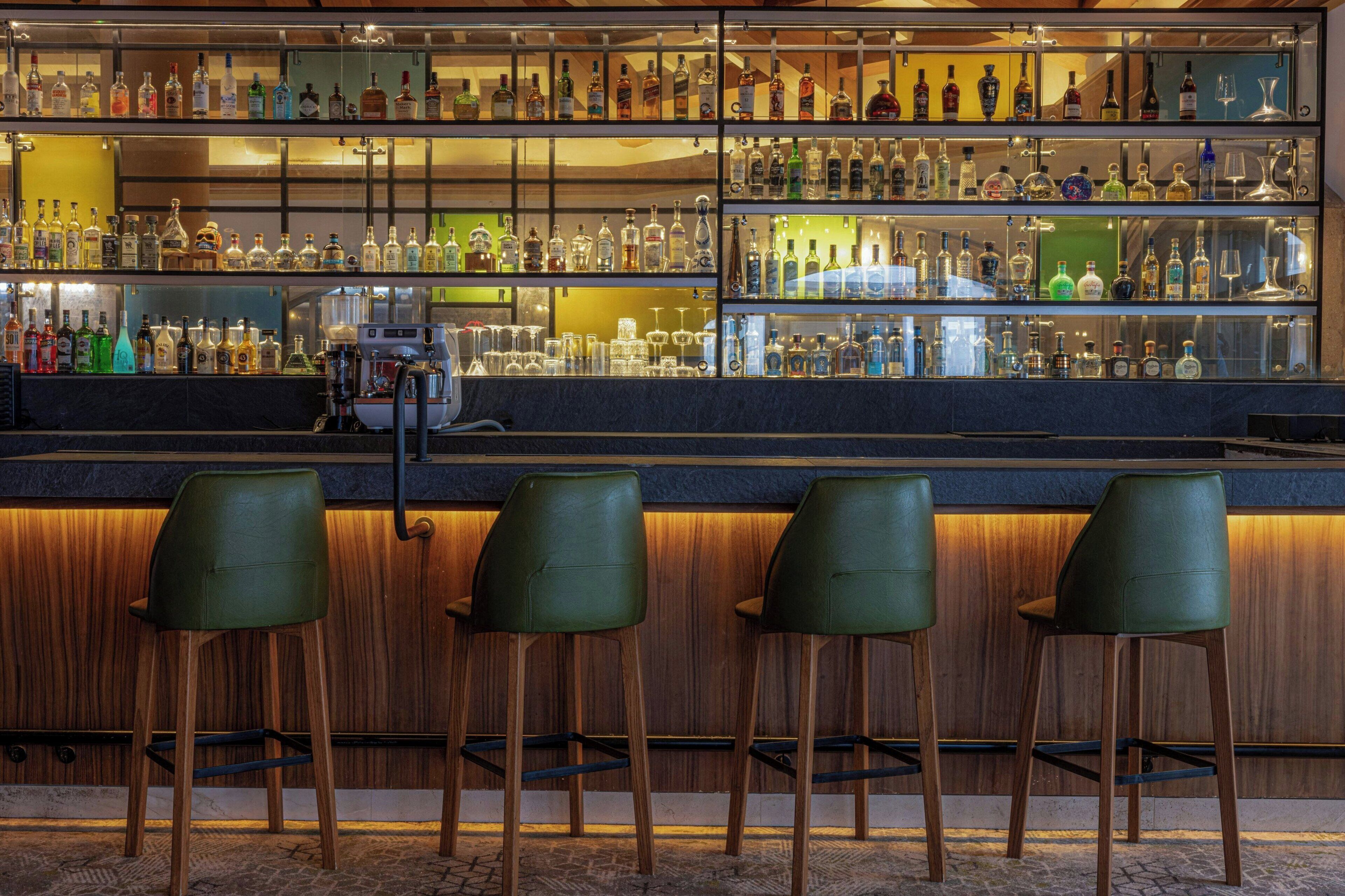 Uma área de bar com várias prateleiras de garrafas e copos, balcão de madeira e três banquetas verdes.