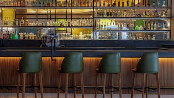 Uma área de bar com várias prateleiras de garrafas e copos, balcão de madeira e três banquetas verdes.