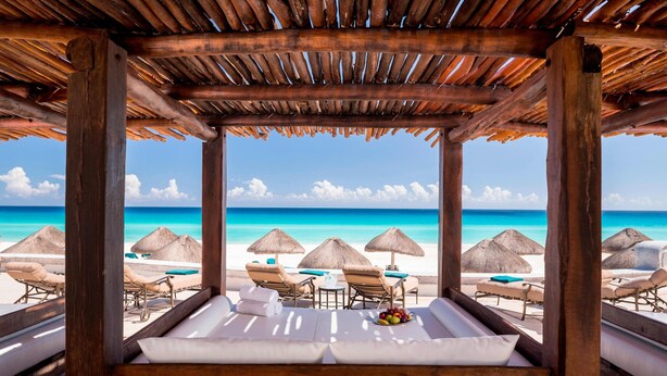 Una cabaña de playa con techo de madera, asientos acolchados blancos y vista al océano.