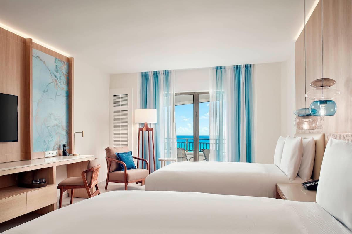 A hotel room with two beds, a desk, a chair, a TV, and a view of the ocean.