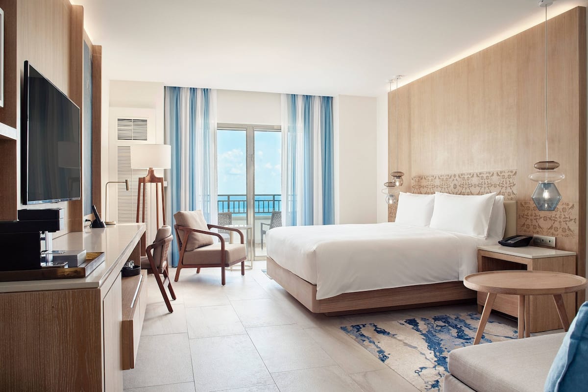 A modern hotel room with a large bed, a desk with a flat-screen TV, a seating area with chairs, and a view of the outdoors through a large window.