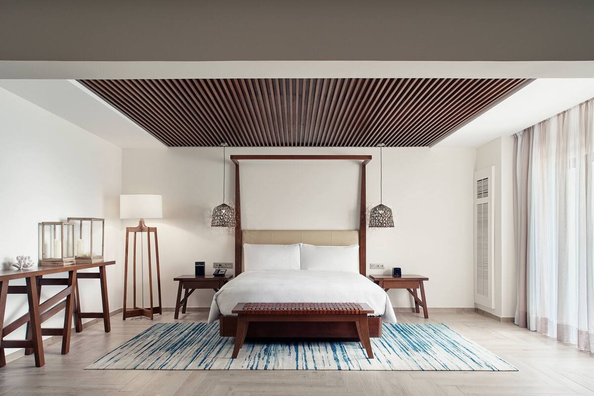 A modern hotel room with a large bed, wooden headboard, bedside tables, a desk, and a lamp.