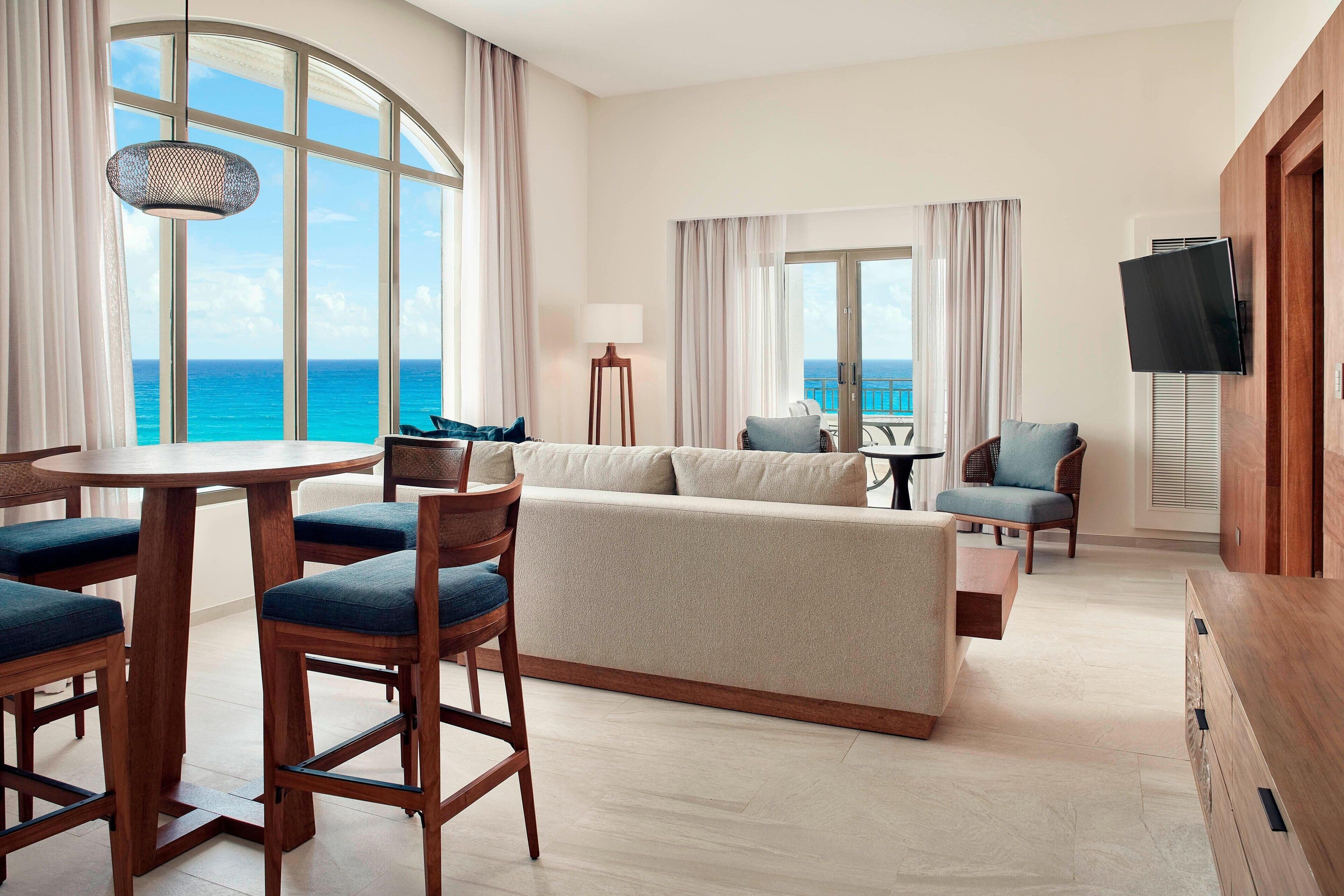 A hotel room with a large window offering a view of the ocean, a sofa, a dining table with chairs, and a television mounted on the wall.