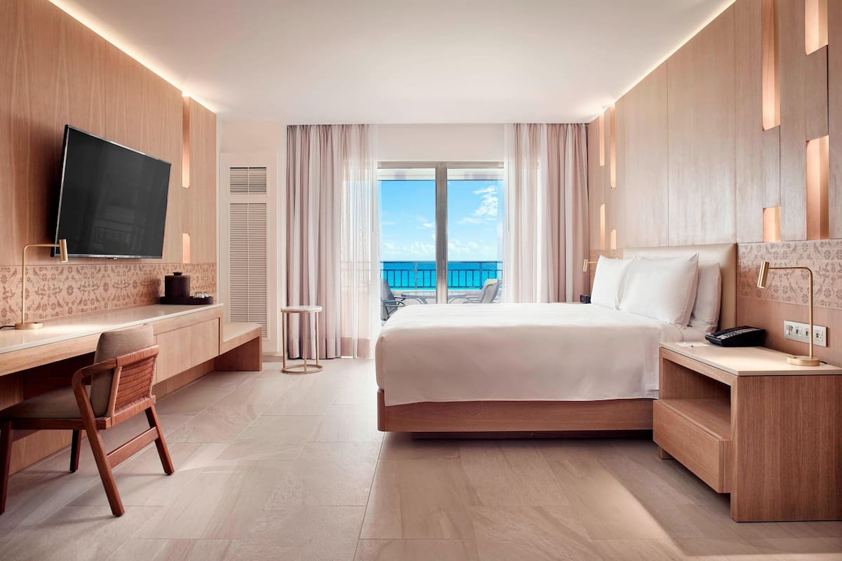 A modern hotel room with a large bed, a desk, a chair, a TV, and a balcony with a view of the sea.