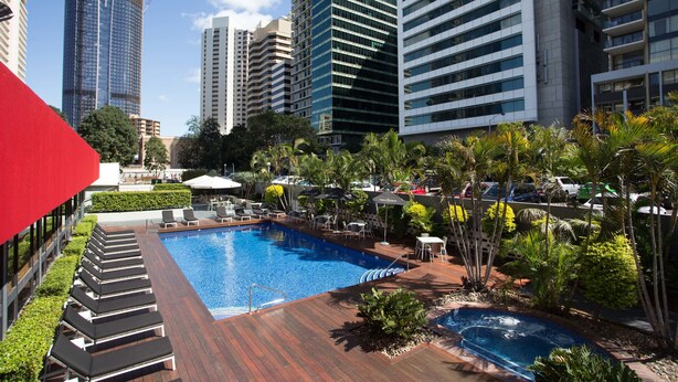 Zona de piscina en la azotea con sillones de descanso y sombrillas, rodeada de edificios altos y palmeras.
