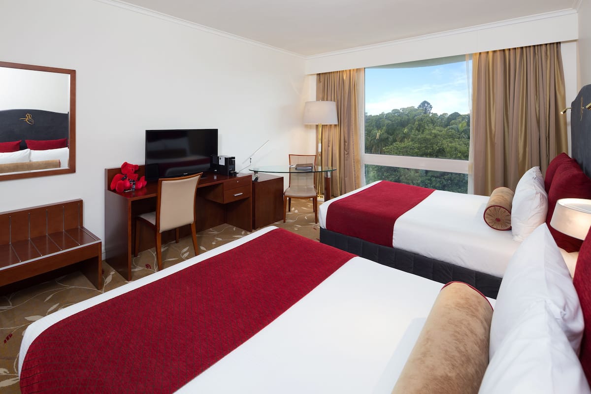A hotel room with two beds, a desk, a TV, and a large window with a view of greenery.