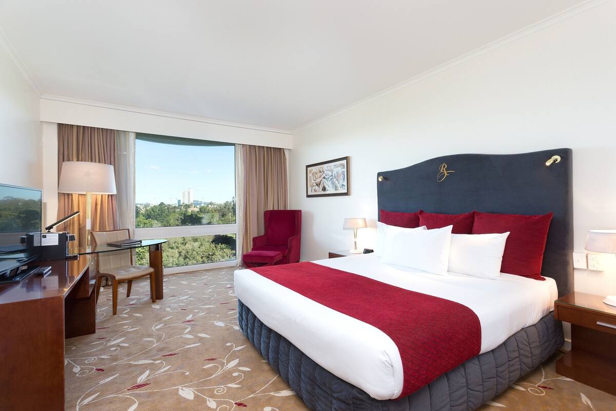 A hotel room with a large bed, a desk, a chair, a TV, and a window with a city view.