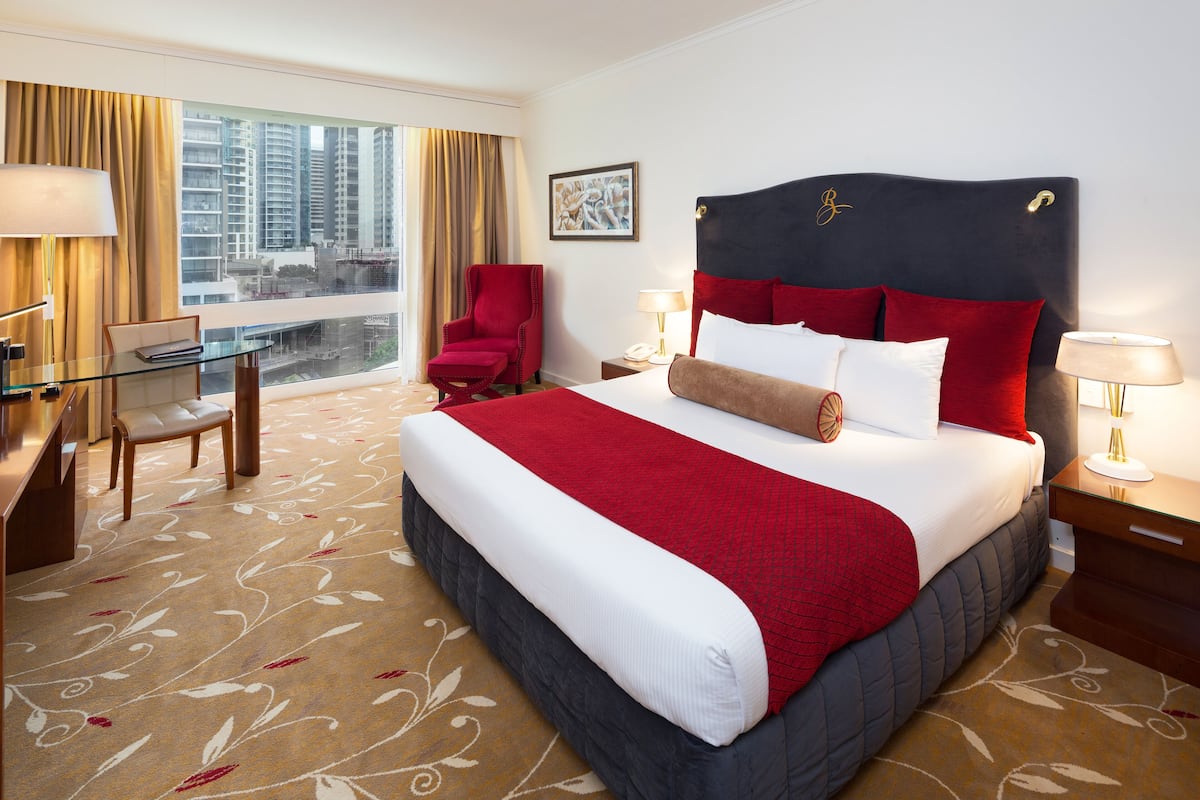 A hotel room with a large bed, red and white bedding, a red armchair, a desk with a chair, and a view of high-rise buildings through the window.