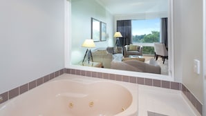 Un baño moderno con una bañera grande, un espejo que refleja un espacio de estar con un sofá y una mesa, y vistas a la vegetación exterior.