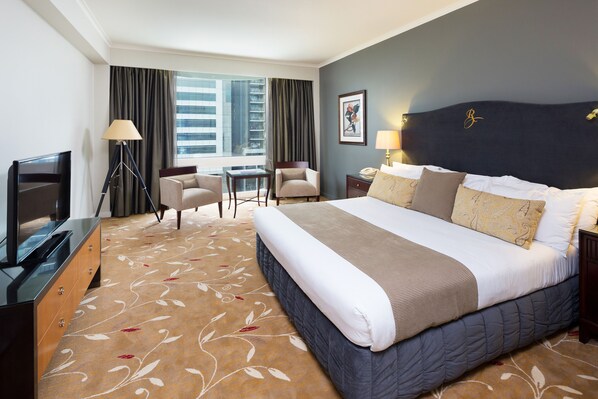 A hotel room with a large bed, a desk, two chairs, a TV, and a view of the city.