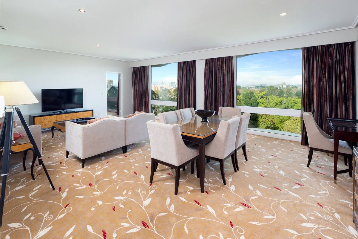 A modern hotel room with a large window, a dining area with a table and chairs, a flat-screen TV, and a sofa.