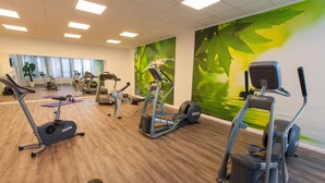 Une salle de sport équipée de divers appareils d’exercice, notamment des tapis de course et des vélos elliptiques, avec un plancher en bois et une fresque représentant un paysage verdoyant sur le mur.