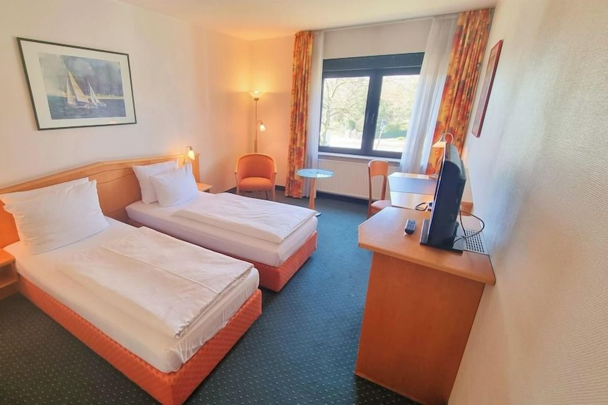 Een hotelkamer met twee bedden, een bureau met televisie, een stoel, een klein tafeltje en een raam met gordijnen.