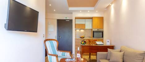 Een moderne hotelkamer met een flatscreen televisie, een houten eettafel met twee stoelen, een bank en een kitchenette op de achtergrond.