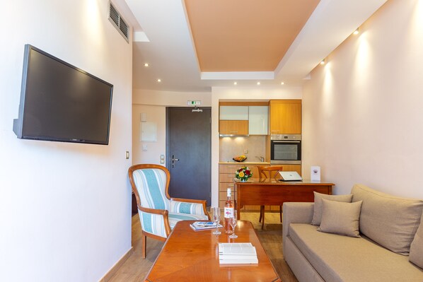 A modern hotel room with a flat-screen TV, a wooden dining table with two chairs, a sofa, and a kitchenette in the background. - Oasis Hotel Apartments (Glyfada)