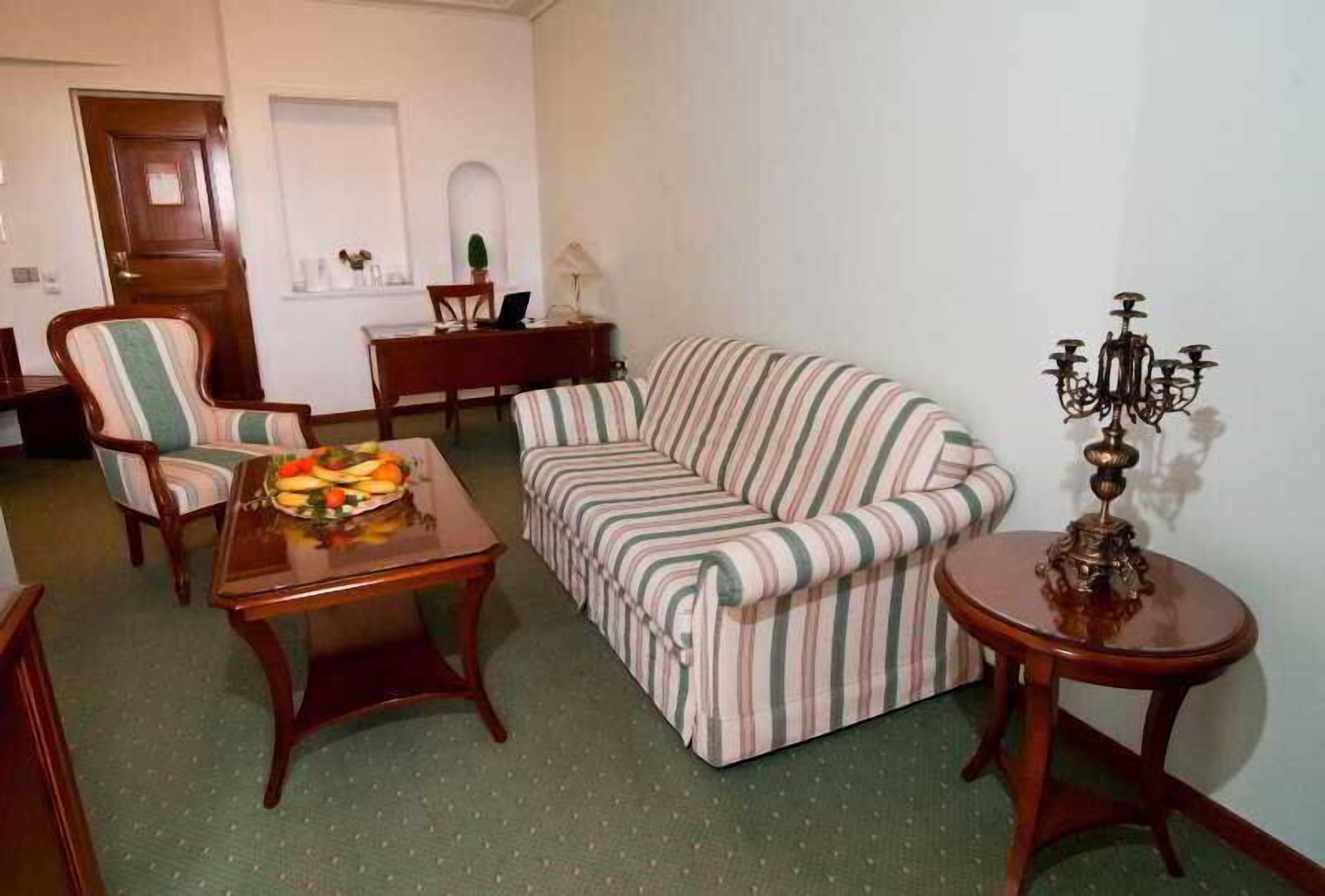 Ein Wohnzimmer mit einem gestreiften Sofa, einem Esstisch mit Obst, einem Sessel, einem kleinen Beistelltisch und einer Chandelier.