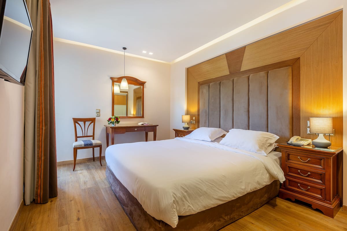 A hotel room with a large bed, a wooden headboard, a desk with a chair, a mirror, and a bedside table with a lamp.