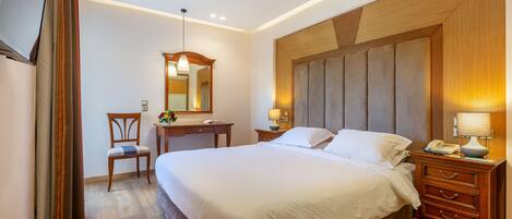 A hotel room with a large bed, a wooden headboard, a desk with a chair, a mirror, and a bedside table with a lamp.