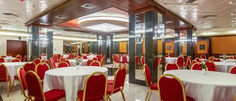 A banquet hall with round tables and red chairs, featuring a high ceiling with recessed lighting and a central chandelier.