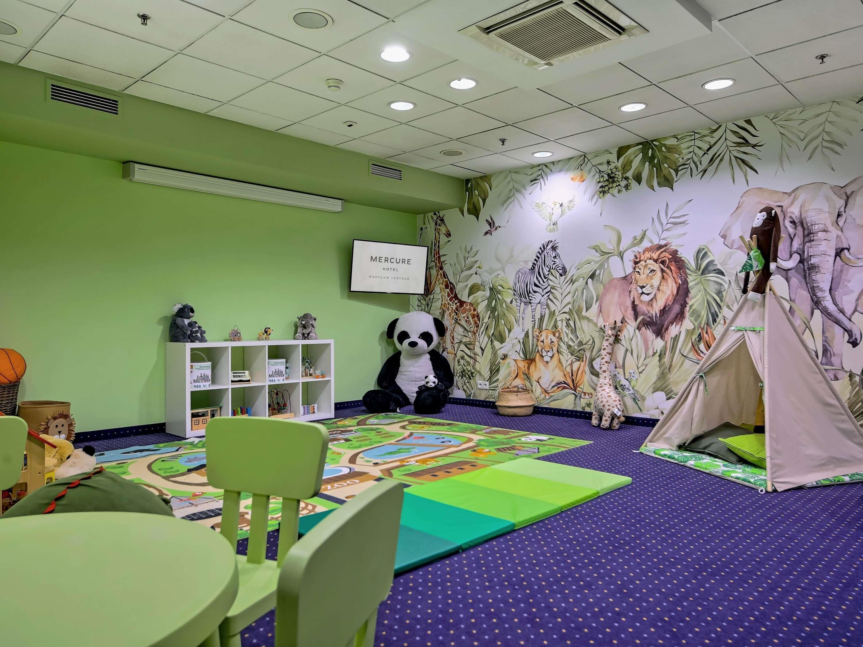 A children's playroom with a green wall, a mural of animals, a white shelf with toys, and a tent on the floor.