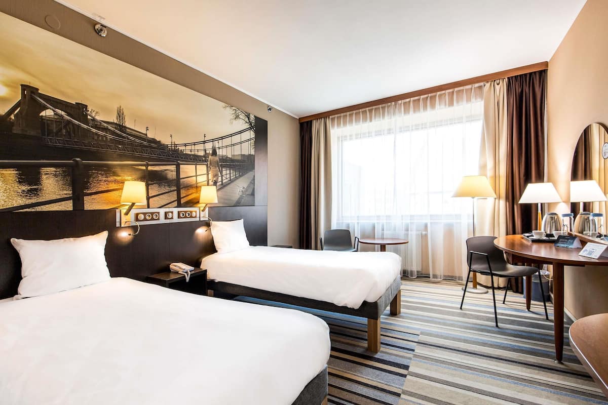 A hotel room with two beds, a desk with a chair, a small table, and a large window with curtains.