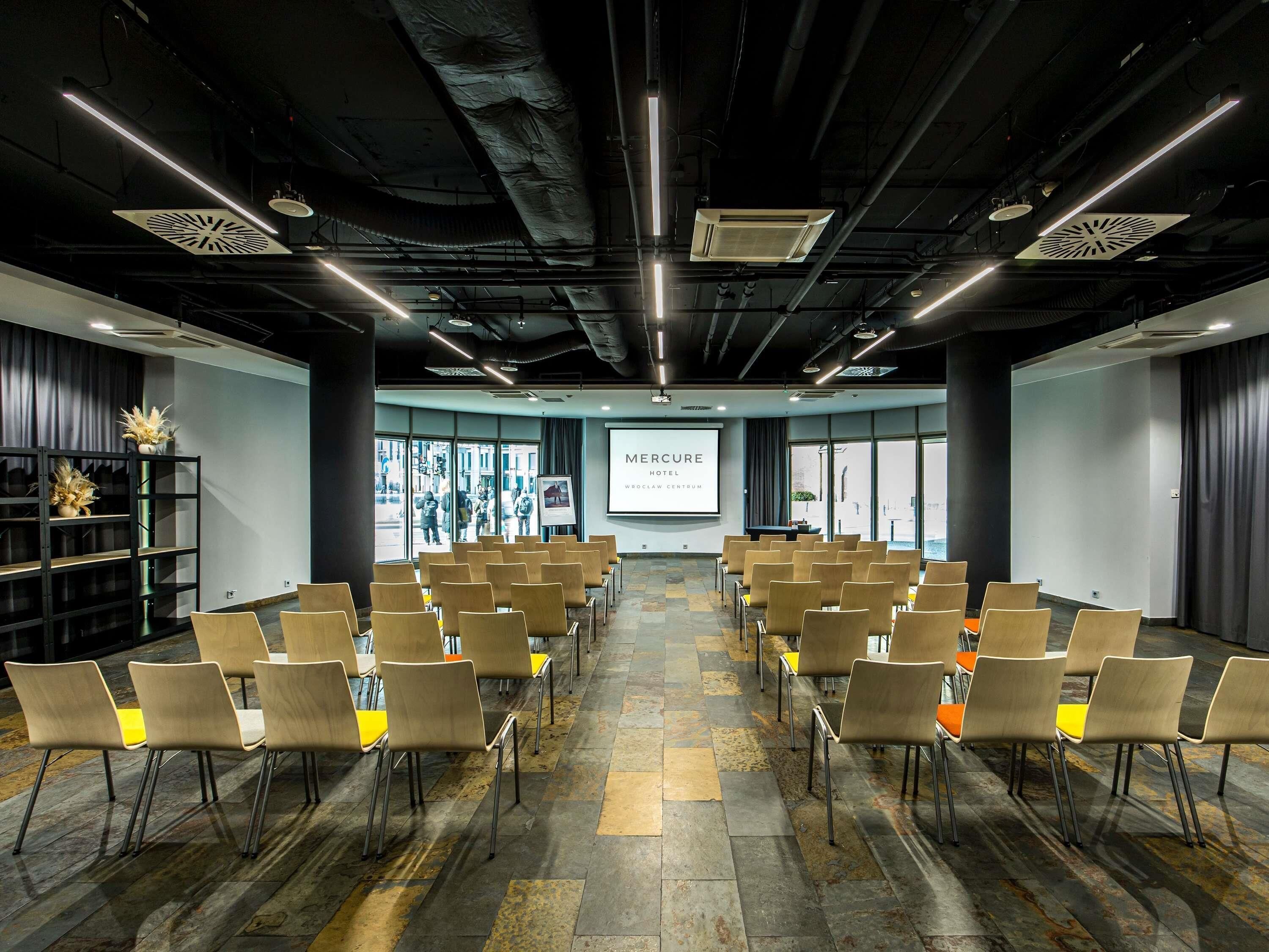 A conference room with rows of chairs, a projector screen, and a large window with a view of the street.
