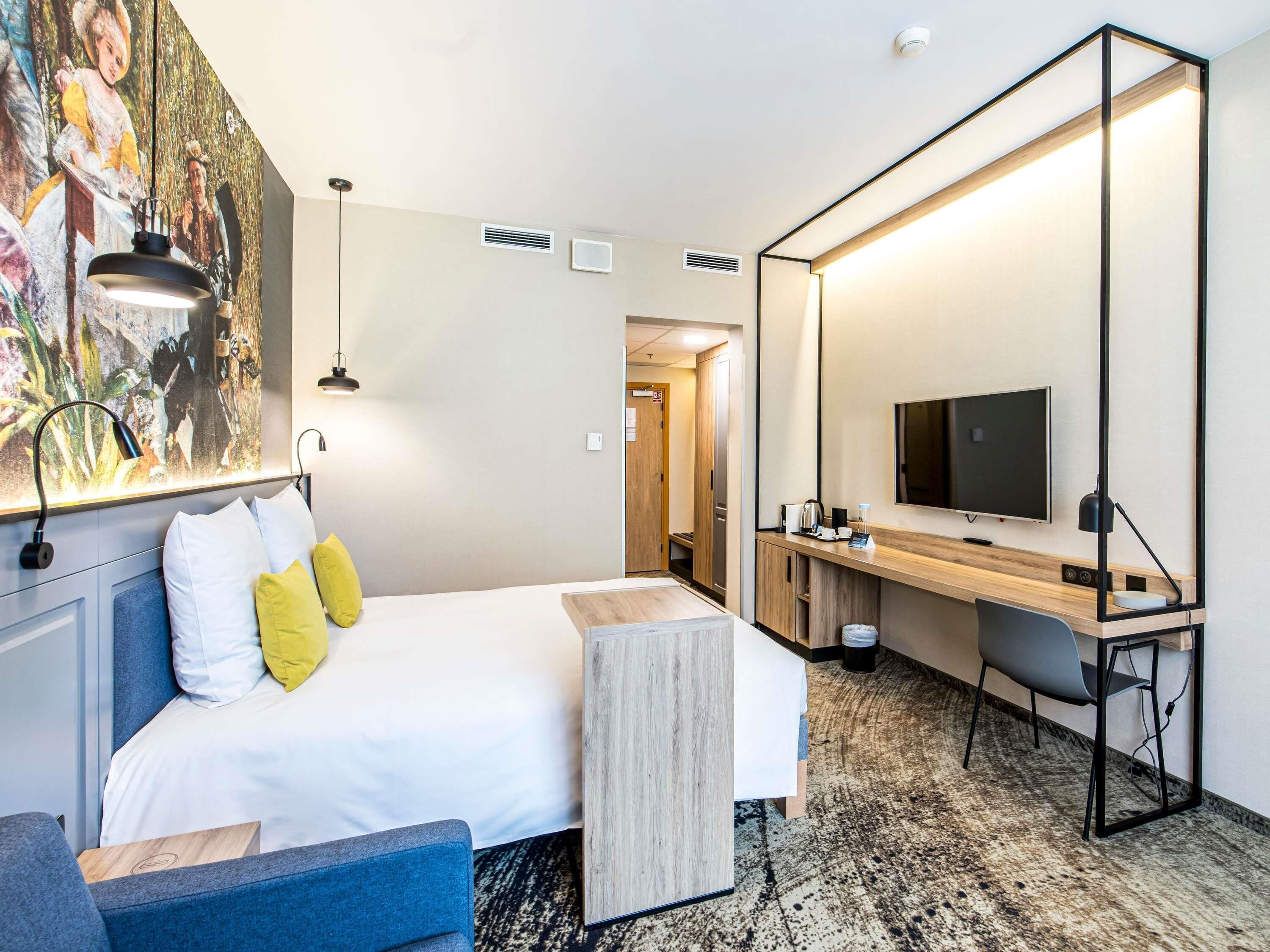 A hotel room with a bed, a desk, a TV, and a large painting on the wall.