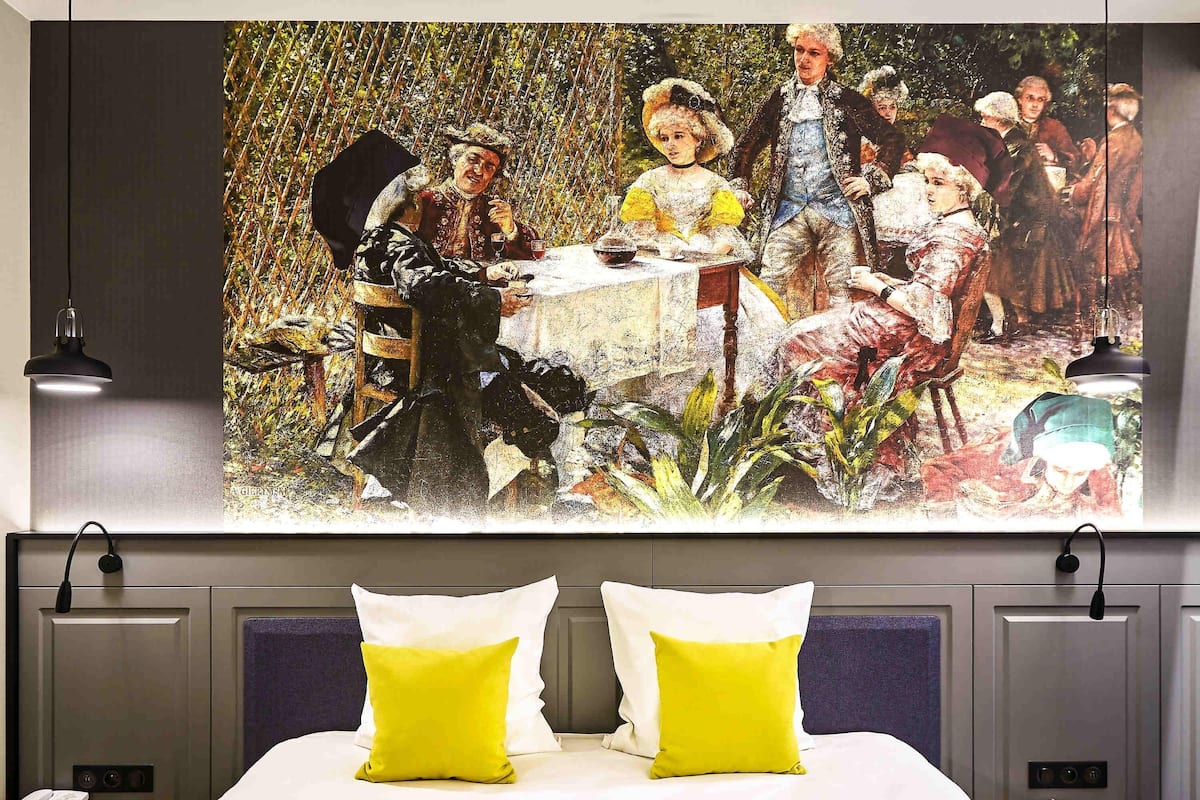 A hotel room with a bed, two bedside lamps, a nightstand with a phone and remote, and a large painting above the bed.