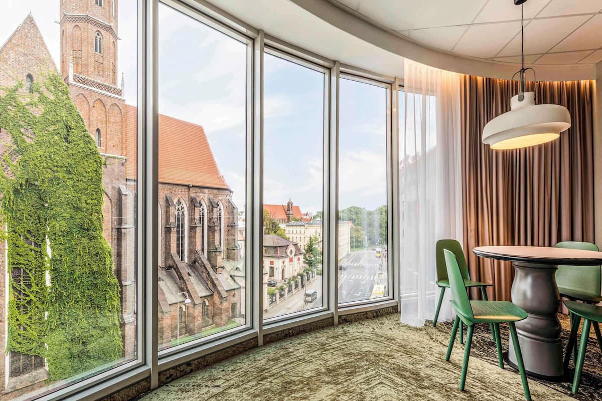 Una habitación con ventanas amplias, una mesa de comedor, sillas, una lámpara colgante y vistas a un edificio histórico.