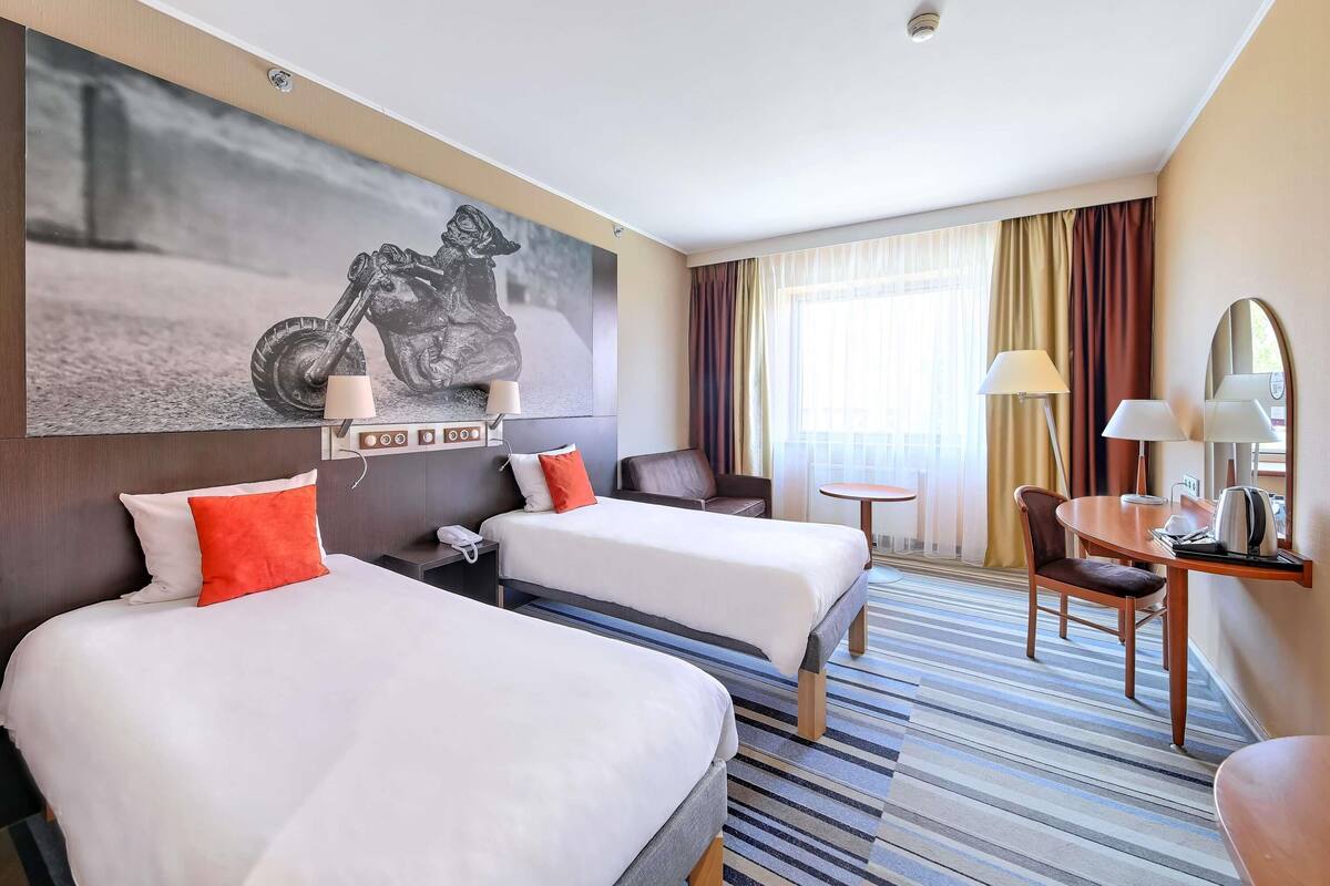 Habitación de hotel con dos camas, un sofá, una mesita y una silla. Se observa una obra de arte grande en la pared con una motocicleta, una ventana con cortinas y una lámpara en el techo.