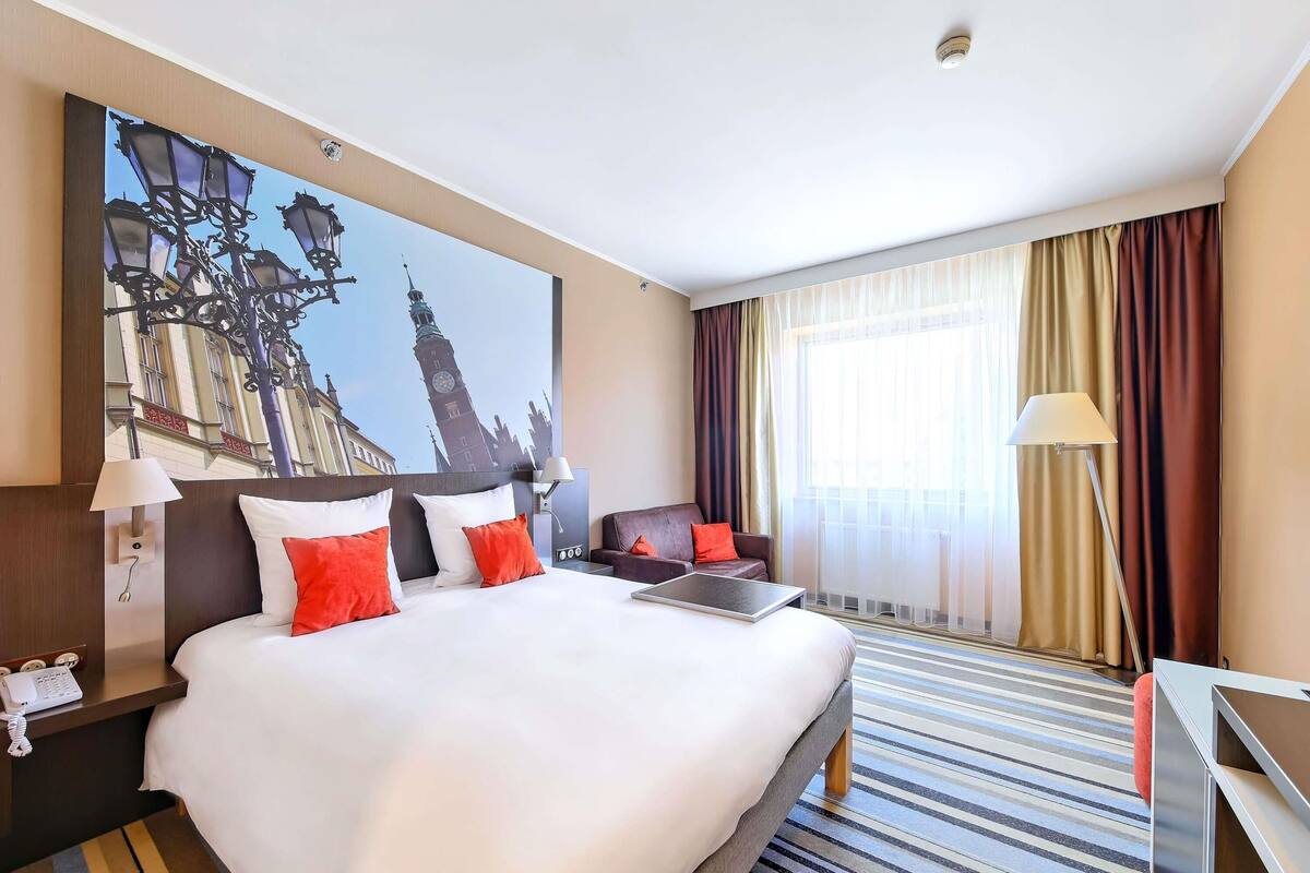 Habitación de hotel con una cama grande, un escritorio, una silla, una lámpara, un cuadro de una torre y una ventana con cortinas.