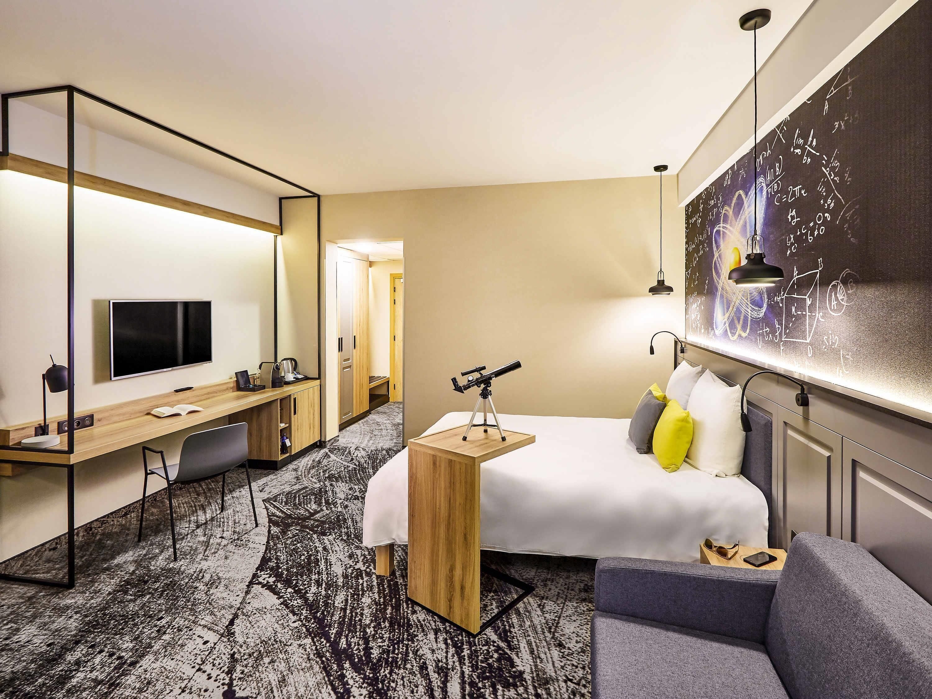 Habitación de hotel con cama, escritorio con televisor, telescopio y pizarra con ecuaciones.