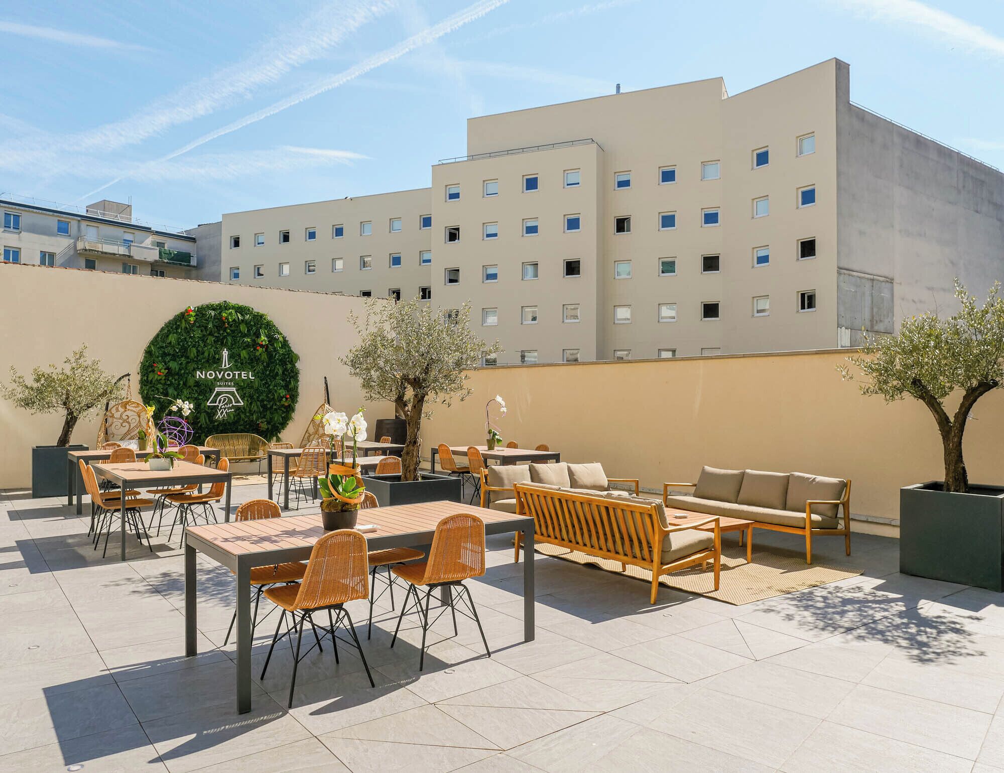 An outdoor seating area with tables and chairs, a decorative wall with greenery, and a building in the background.