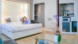 Una habitación de hotel moderna con una cama grande, una mesita de centro de cristal, una pequeña cocina y una planta en maceta.