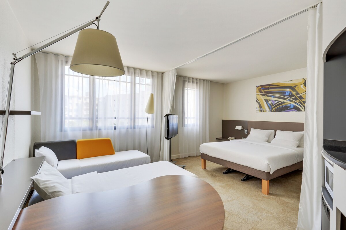 A hotel room with two beds, a desk, a TV, and a large window with curtains.