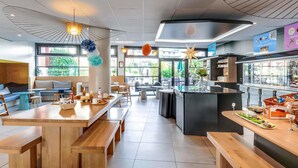 Un café moderno con mesas y bancos de madera, una isla central con una máquina de café y vistas a zonas verdes al exterior.