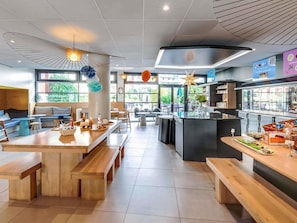 A modern cafe with wooden tables and benches, a central island with a coffee machine, and a view of greenery outside.