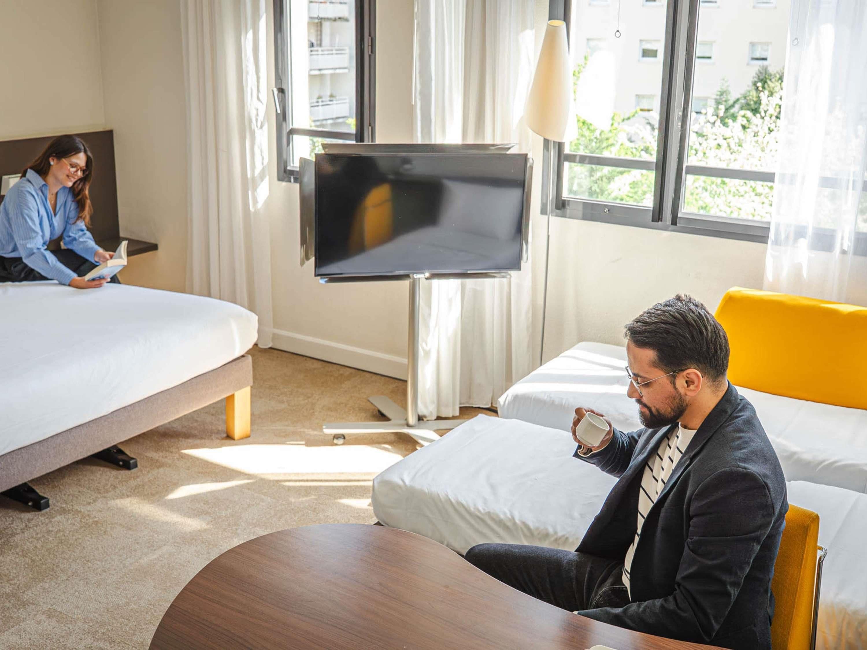 Quarto de hotel com cama, televisão, escrivaninha e janela com vista para edifícios e áreas verdes.