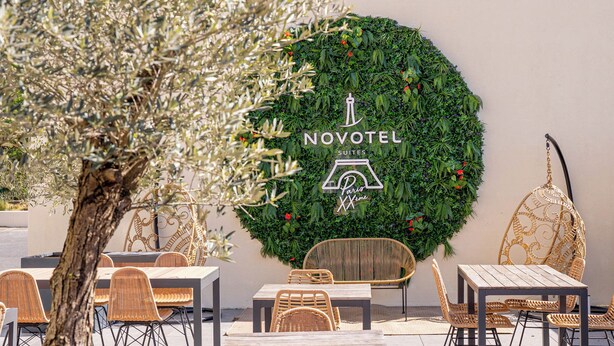 Un área de estar al aire libre con sillas y mesas de mimbre, un muro decorativo con follaje y el logotipo de Novotel, y un árbol al fondo.