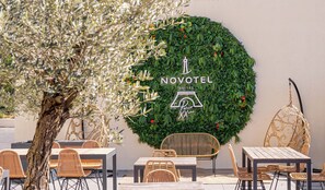 Un área de estar al aire libre con sillas y mesas de mimbre, un muro decorativo con follaje y el logotipo de Novotel, y un árbol al fondo.