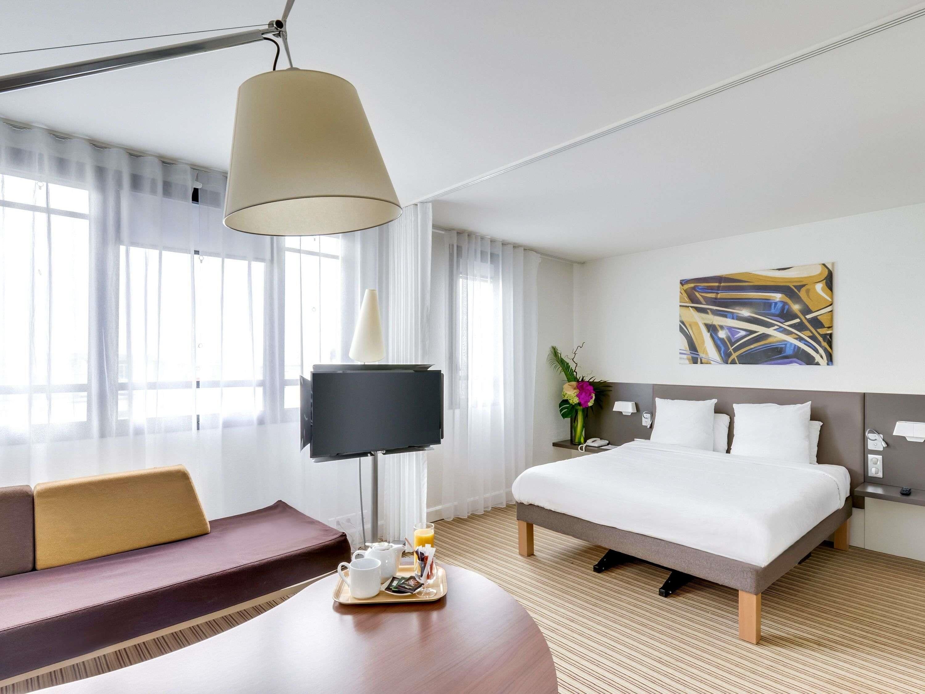A hotel room with a bed, a sofa, a table with a tray of breakfast items, a lamp, and a painting on the wall.