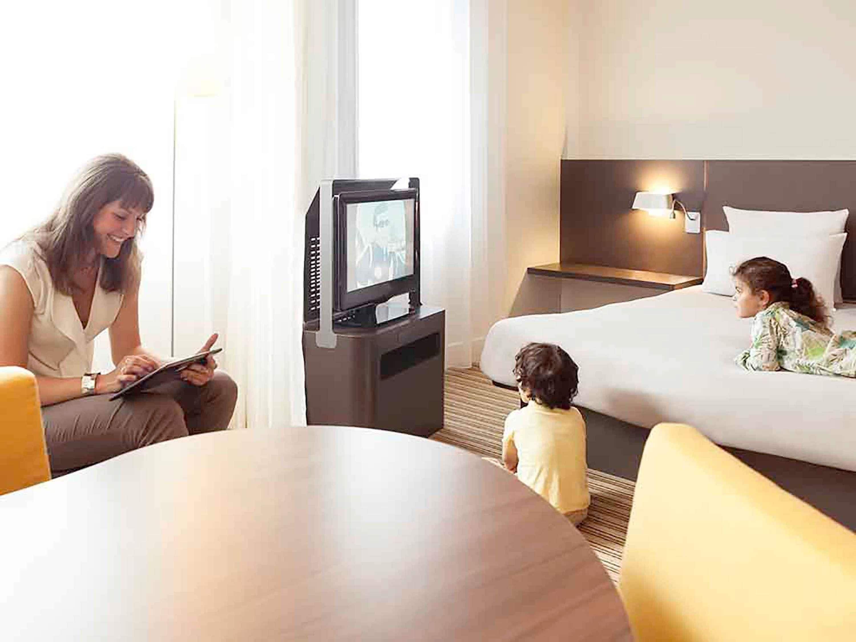 A hotel room with a bed, a television, a table, and a child.