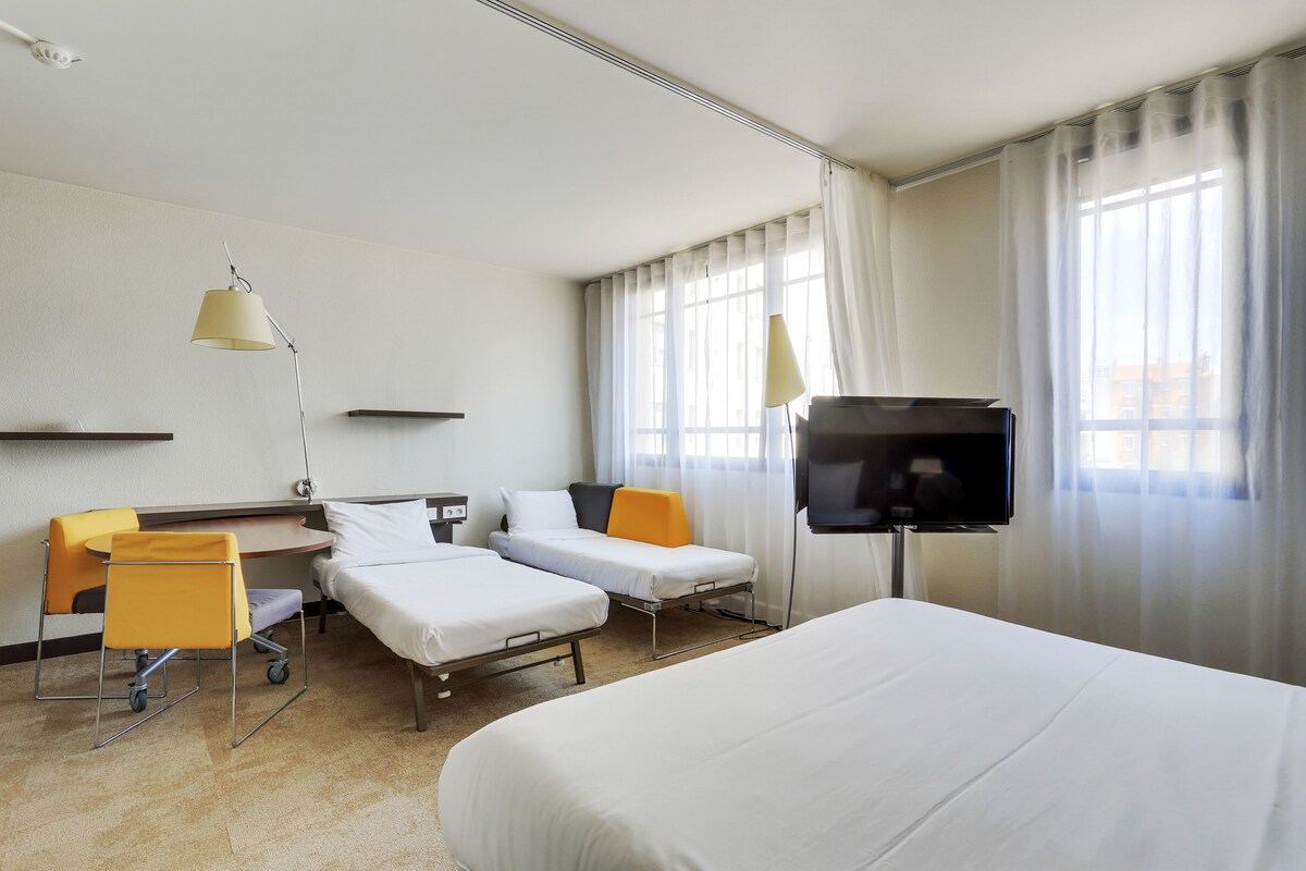 A hotel room with two single beds, a desk with a chair, a TV, and a window with curtains.