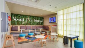 A hotel lobby with a green wall, seating area, tables, and a television. - Novotel Suites Paris Montreuil Vincennes (Paris)