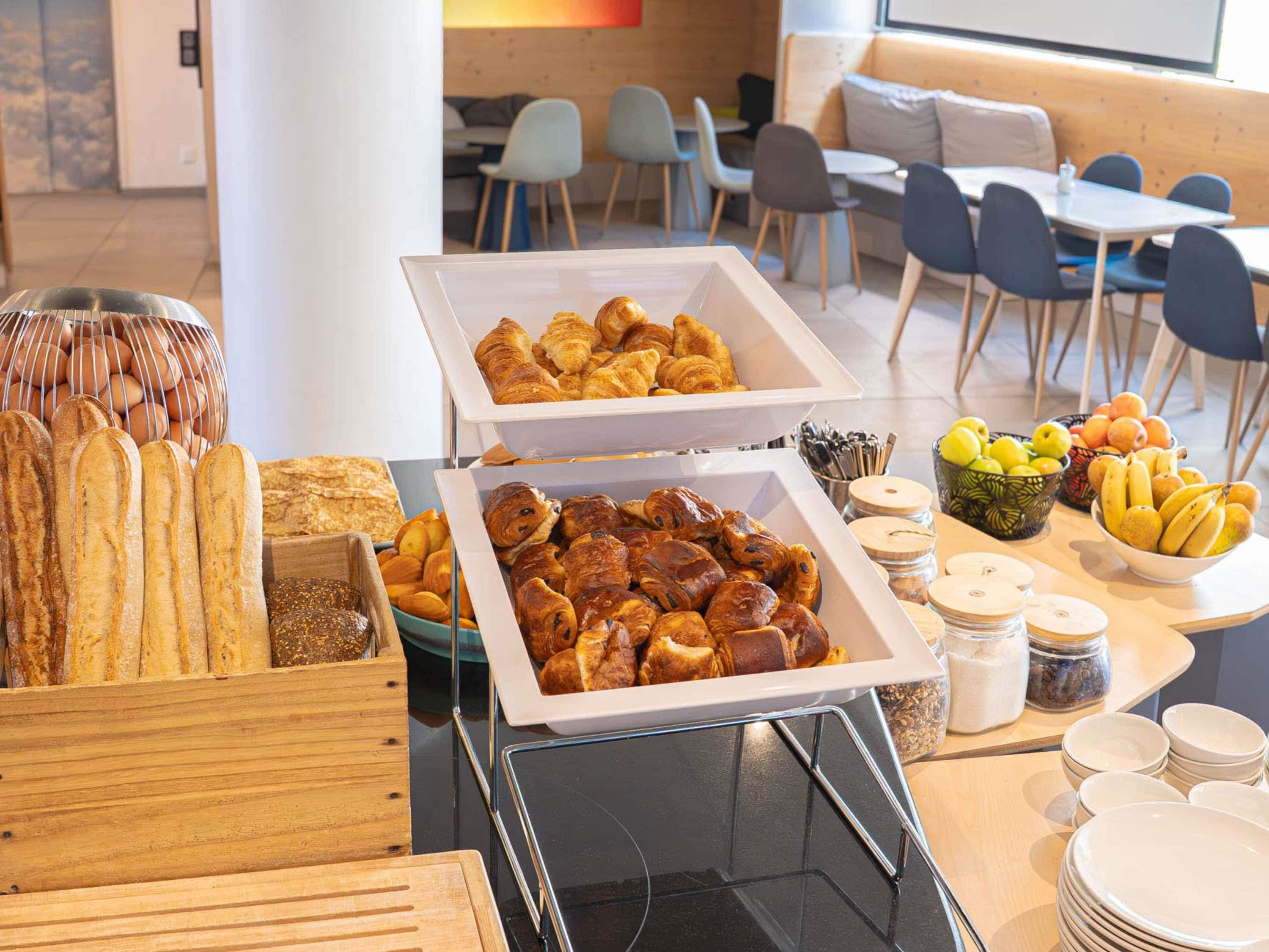 Montaje de bufé con pan, bollería y frutas en un comedor moderno con sillas y mesas.