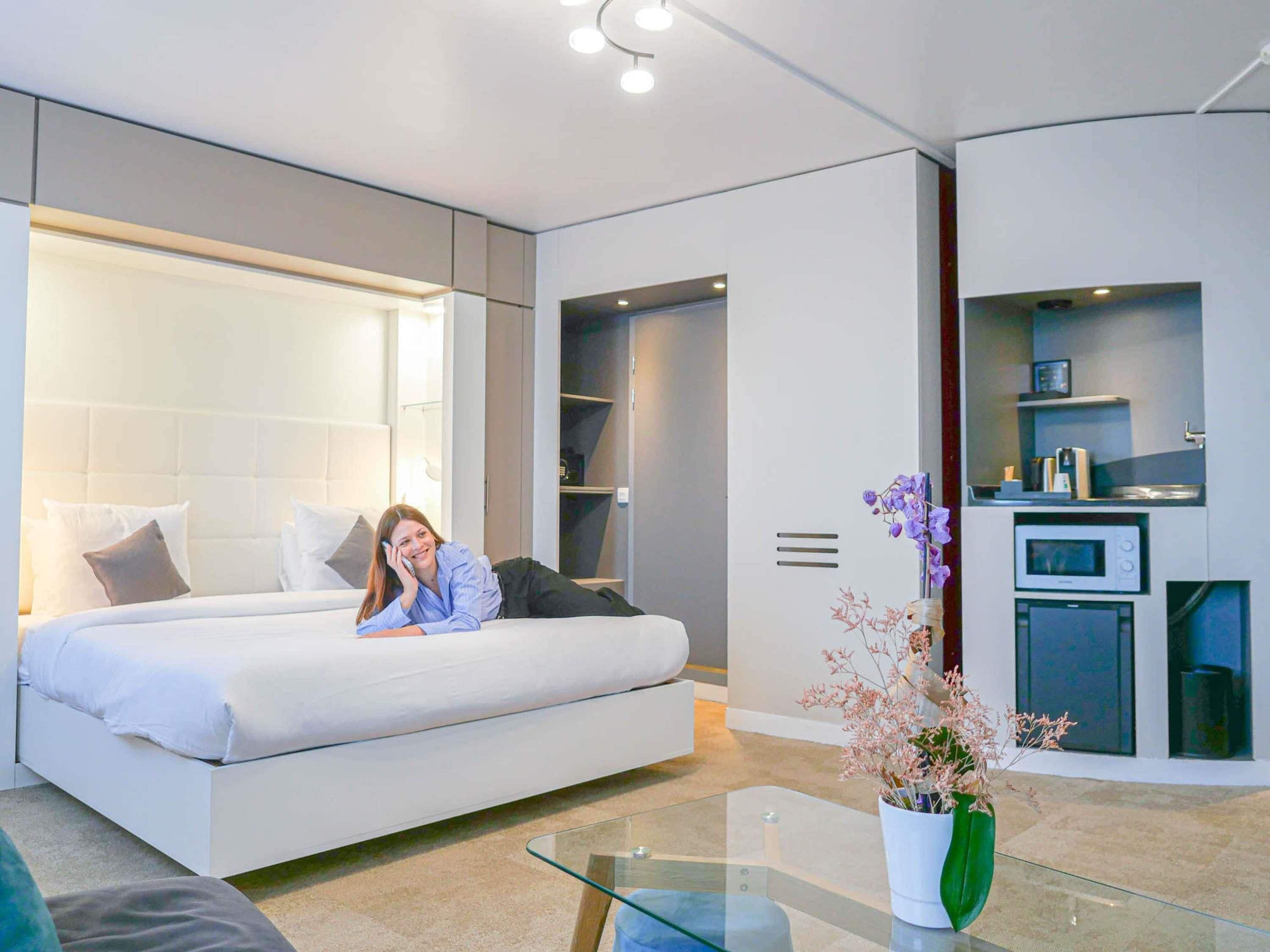A modern hotel room with a large bed, a glass coffee table, a small kitchen area, and a potted plant.