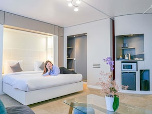 Une chambre d’hôtel moderne avec un grand lit, une table basse en verre, un petit coin cuisine et une plante en pot.