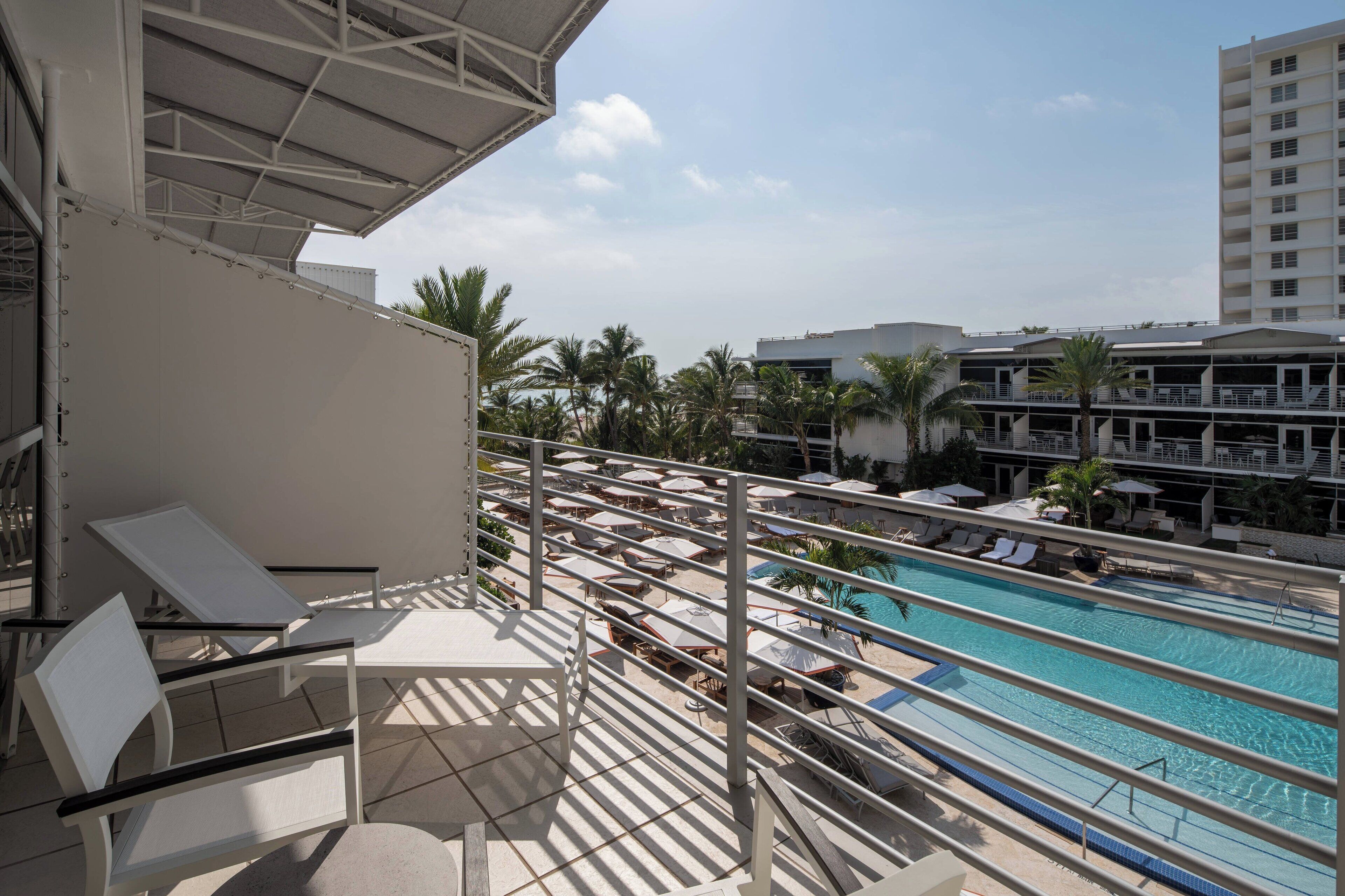A balcony with white chairs and a pool area with lounge chairs and umbrellas.
