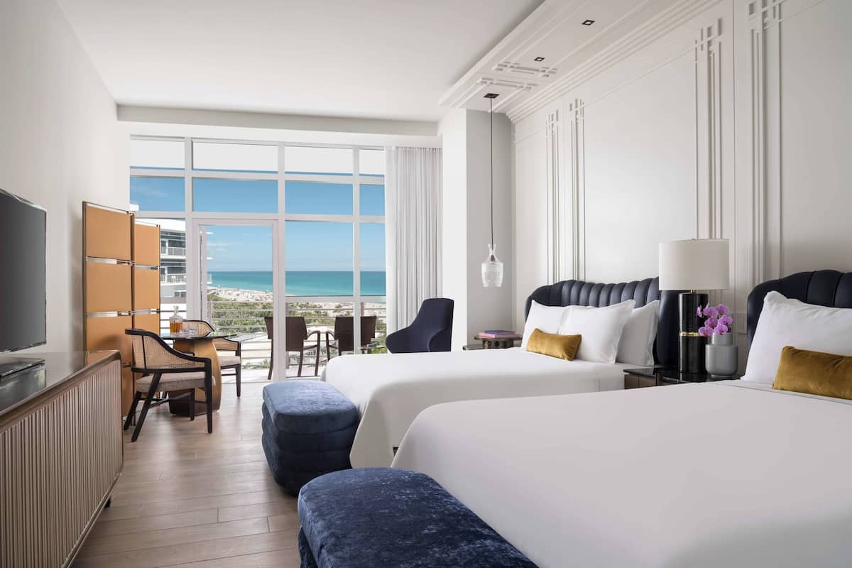 A hotel room with two beds, a TV, a desk, and a view of the ocean.